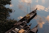 Wooden boats are moored on a calm water surface, reflecting a cloud-dappled sky. Yellow crates and other items are visible on the boats. Lush greenery lines the edge of the water, adding a natural border.