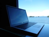 A laptop is placed on a reflective surface, with a view of a serene body of water and small islands in the background. The sky is bright blue with scattered clouds, contributing to a tranquil setting.