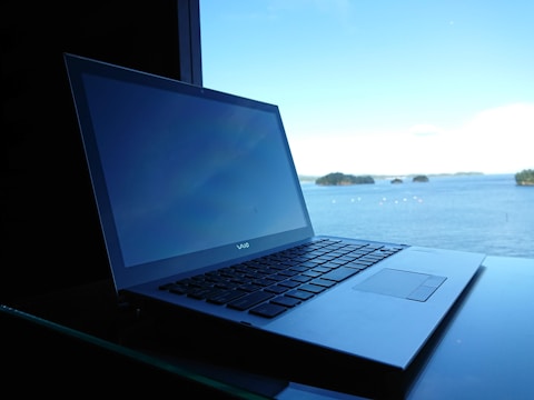 A laptop is placed on a reflective surface, with a view of a serene body of water and small islands in the background. The sky is bright blue with scattered clouds, contributing to a tranquil setting.