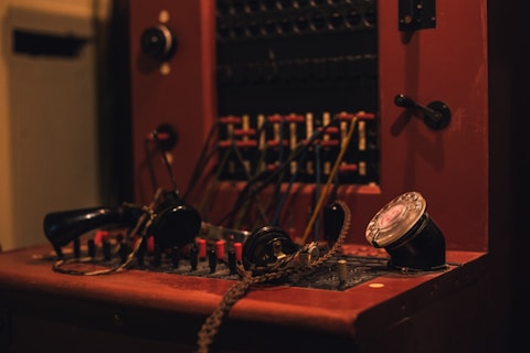 A vintage telephone switchboard with numerous wires, a rotary dial, and a handset. The setup includes various plugs and connectors, all displayed on a wooden panel.