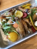 A metal tray containing crispy baked chicken wings garnished with sliced green onions and accompanied by colorful bell pepper slices, including red and yellow, as well as bits of cauliflower.