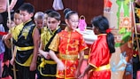 A group of children stands together wearing traditional Chinese clothing in bright colors. Most of them are dressed in black and gold outfits, with one child in a prominent red outfit featuring a gold dragon pattern. They are gathered in front of a colorful backdrop depicting traditional Chinese architecture and motifs.