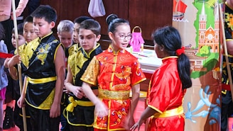 A group of children stands together wearing traditional Chinese clothing in bright colors. Most of them are dressed in black and gold outfits, with one child in a prominent red outfit featuring a gold dragon pattern. They are gathered in front of a colorful backdrop depicting traditional Chinese architecture and motifs.