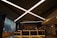 A modern reception area with a sleek, dark ceiling featuring intersecting linear light fixtures. The reception desk is staffed by three individuals and has a vase with brightly colored flowers as a centerpiece. There is a roped-off area in front of the desk and large windows on the left side of the space. Various office equipment and decor can be seen on and behind the desk.
