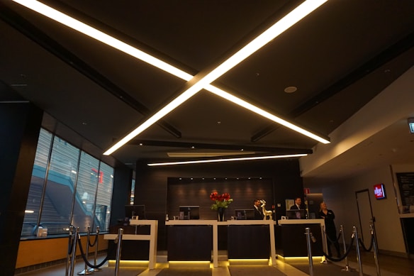 A modern reception area with a sleek, dark ceiling featuring intersecting linear light fixtures. The reception desk is staffed by three individuals and has a vase with brightly colored flowers as a centerpiece. There is a roped-off area in front of the desk and large windows on the left side of the space. Various office equipment and decor can be seen on and behind the desk.