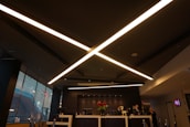 A modern, dimly lit interior with a sleek design dominated by large, criss-crossing ceiling lights. The reception area features a desk with red flowers and several people behind it. Large windows on the side let in some natural light, and the atmosphere is professional.