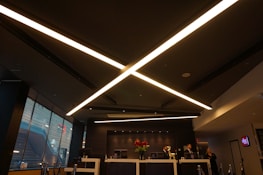 A modern, dimly lit interior with a sleek design dominated by large, criss-crossing ceiling lights. The reception area features a desk with red flowers and several people behind it. Large windows on the side let in some natural light, and the atmosphere is professional.