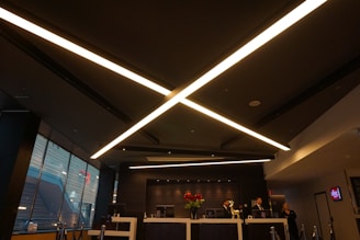 A modern, dimly lit interior with a sleek design dominated by large, criss-crossing ceiling lights. The reception area features a desk with red flowers and several people behind it. Large windows on the side let in some natural light, and the atmosphere is professional.