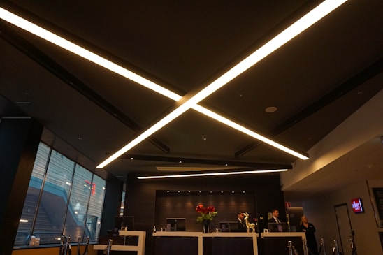 A modern, dimly lit interior with a sleek design dominated by large, criss-crossing ceiling lights. The reception area features a desk with red flowers and several people behind it. Large windows on the side let in some natural light, and the atmosphere is professional.