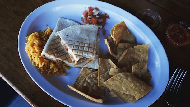 A white oval plate holds a serving of yellow rice, a folded quesadilla with grilled marks, a side of fresh salsa, and a portion of crispy tortilla chips. The plate is set on a wooden table with a fork visible to the side.