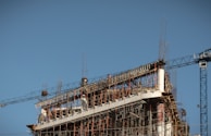 A partially constructed building with an exposed framework of steel beams and scaffolding. A large construction crane is visible on the right, and the sky is clear and blue.