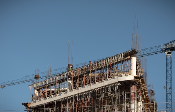 A partially constructed building with an exposed framework of steel beams and scaffolding. A large construction crane is visible on the right, and the sky is clear and blue.
