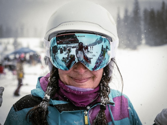 A person wearing a white helmet and large reflective ski goggles stands in a snowy environment. The goggles reflect a scene with several people, skis, and equipment in a wintry landscape. The person is dressed in winter clothing with a noticeable blue and purple color scheme, and there is snow on their hair and attire.