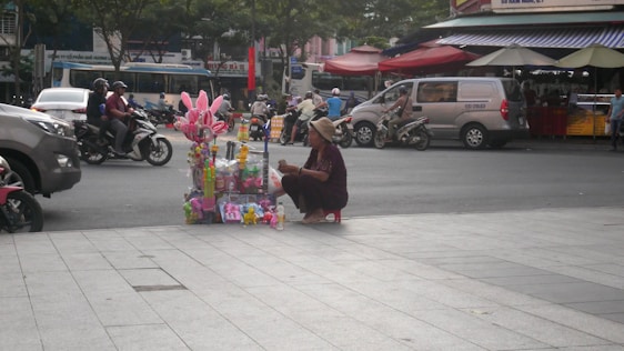 A street vendor is sitting on a small stool next to a variety of colorful toys displayed on a street corner. Motorcycles and cars pass by on the road, and there are shops and street stalls with awnings in the background.