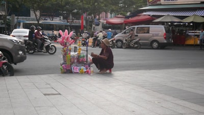 A street vendor is sitting on a small stool next to a variety of colorful toys displayed on a street corner. Motorcycles and cars pass by on the road, and there are shops and street stalls with awnings in the background.