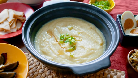 A bowl of creamy porridge-like dish garnished with chopped green onions, placed on a woven mat. Surrounding the main dish, there are arranged plates containing sliced ingredients, boiled eggs, and other condiments. The setting has a homely and inviting appearance with a red tablecloth.