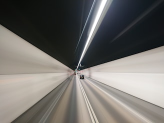 A long tunnel with two lanes and modern lighting, presenting a sense of motion blur. A car can be seen driving in the right lane, heading towards the distant vanishing point in the tunnel. The walls are uniformly light-colored with dark ceiling and bright, linear lights running along the central line above.