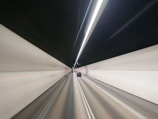 A long tunnel with two lanes and modern lighting, presenting a sense of motion blur. A car can be seen driving in the right lane, heading towards the distant vanishing point in the tunnel. The walls are uniformly light-colored with dark ceiling and bright, linear lights running along the central line above.