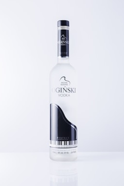 A vodka bottle featuring a sleek design with a black and white grand piano image. The label includes text in an elegant font and highlights the name 'OGINSKI VODKA'. The bottle is clear and sits against a plain, light background.