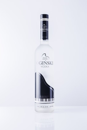 A vodka bottle featuring a sleek design with a black and white grand piano image. The label includes text in an elegant font and highlights the name 'OGINSKI VODKA'. The bottle is clear and sits against a plain, light background.