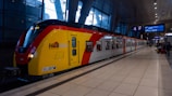 A modern, colorful train is stationed at a platform in an indoor railway station. The train is predominantly yellow with red and black accents and features the logo of HLB and other signage. Several passengers can be seen on the platform, some with luggage, waiting or walking by. The station is spacious with a high ceiling and large windows that allow natural light in.