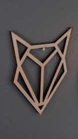 A geometric wooden wall art piece resembling the shape of an animal face, possibly a fox, made from intersecting lines. It is mounted on a smooth, dark gray wall using a small metal nail.