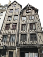A pair of historic timber-framed buildings with detailed wooden beams and small-paned windows situated closely together. The structures feature multiple stories with gabled roofs and are made of a mix of stone and wood, showcasing traditional architectural techniques.