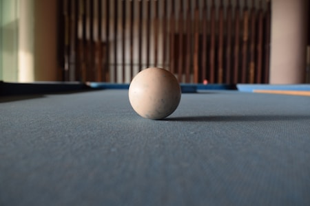 A solitary billiard ball rests on a pool table with a focus on the texture of the felt. The background features vertical wooden bars, creating a sense of depth.