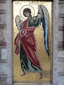 A detailed mosaic artwork depicting an angel with a halo, wearing a red and green robe. The angel is holding a staff and has wings with shades of blue. The background is made up of small golden tiles, creating a shimmering effect. Greek text is visible at the top and bottom of the mosaic, which is framed by a stone border.