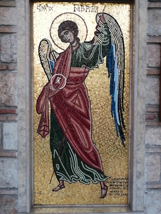 A detailed mosaic artwork depicting an angel with a halo, wearing a red and green robe. The angel is holding a staff and has wings with shades of blue. The background is made up of small golden tiles, creating a shimmering effect. Greek text is visible at the top and bottom of the mosaic, which is framed by a stone border.