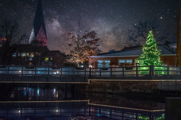 A serene night scene with a beautifully lit Christmas tree glowing in green beside a calm body of water. A tall church steeple rises in the background against a sky filled with stars and the Milky Way. Reflections of the tree and nearby buildings are visible on the water surface.