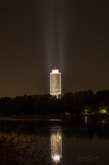 A tall, illuminated tower stands at night, surrounded by trees and casting a bright reflection on a calm body of water. The tower emits beams of light from the top, creating a striking visual effect against the dark sky.