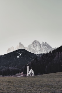 A small church with a tall steeple stands in an open grassy field surrounded by dense dark green forests. In the background, large, rugged mountains covered in a light dusting of snow rise into a pale sky.