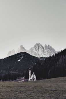 A small church with a tall steeple stands in an open grassy field surrounded by dense dark green forests. In the background, large, rugged mountains covered in a light dusting of snow rise into a pale sky.