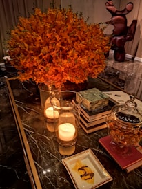 A luxurious interior scene with a large bouquet of vibrant orange flowers, two burning candles in glass holders, a stack of books, a glass jar containing a snack, and a decorative sculpture in the background. The setting conveys a warm and elegant atmosphere.