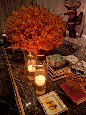 A luxurious interior scene with a large bouquet of vibrant orange flowers, two burning candles in glass holders, a stack of books, a glass jar containing a snack, and a decorative sculpture in the background. The setting conveys a warm and elegant atmosphere.
