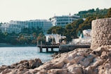 A coastal scene featuring rocky shorelines in the foreground with clear blue waters. Modern resort buildings with balconies rise behind dense green trees. Elegant cabanas with white curtains stand on a dock over the water, and several sun loungers with closed turquoise umbrellas are arranged along the water's edge.