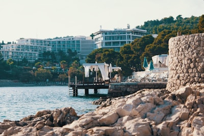 A coastal scene featuring rocky shorelines in the foreground with clear blue waters. Modern resort buildings with balconies rise behind dense green trees. Elegant cabanas with white curtains stand on a dock over the water, and several sun loungers with closed turquoise umbrellas are arranged along the water's edge.