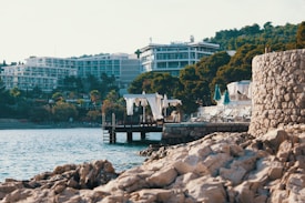 A coastal scene featuring rocky shorelines in the foreground with clear blue waters. Modern resort buildings with balconies rise behind dense green trees. Elegant cabanas with white curtains stand on a dock over the water, and several sun loungers with closed turquoise umbrellas are arranged along the water's edge.
