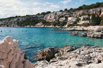 A serene coastal landscape featuring clear blue waters and rocky shores. Along the coastline, several white houses with red roofs are nestled amongst lush green foliage and cliffs. The sky is dotted with a few clouds, creating a tranquil atmosphere.