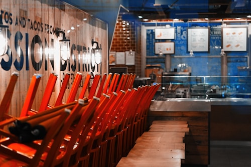 A restaurant interior with orange chairs stacked on tables. The space is dimly lit with warm lighting, showing a wooden wall with large text and light fixtures. A menu is visible in the background, on a bright blue wall above a counter area.