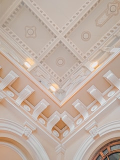 An ornate ceiling with intricate architectural details including moldings, decorative patterns, and recessed lighting. The design features symmetrical geometric shapes and relief decorations, conveying a sense of elegance and classical architecture.