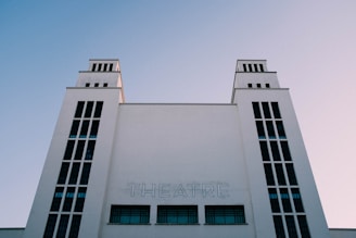 A tall, symmetrical building with a minimalist design stands against a clear sky. The word 'THEATRE' is prominently displayed on the facade. The architecture features vertical windows and two towers.