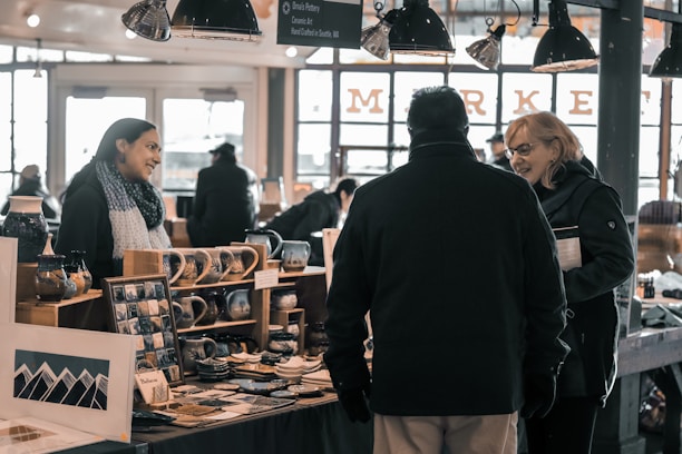 A bustling indoor market stall with pottery items displayed on wooden shelves. Three people are engaged in conversation, with a woman on the left appearing to sell her handmade ceramic products. The atmosphere is lively, with warm lighting illuminating the scene and other market activities visible in the background through large windows.