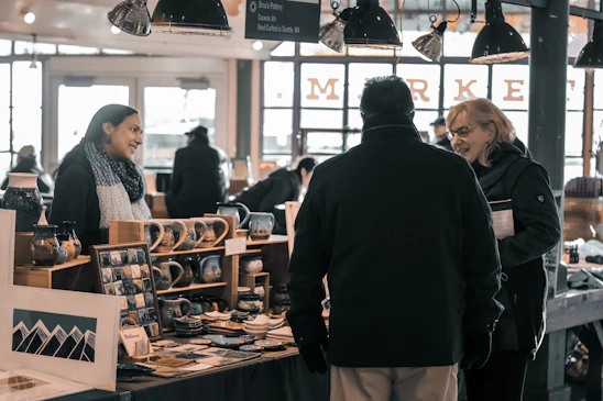 A lively scene of artisans and visitors interacting in a cozy pottery workshop filled with handmade ceramics.