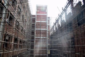 A construction site featuring tall structures covered in scaffolding, with exposed brickwork on the buildings. The scaffolds form a grid pattern around the buildings. The scene appears industrial and shows early stages of development.