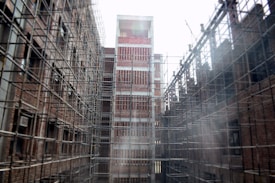 A construction site featuring tall structures covered in scaffolding, with exposed brickwork on the buildings. The scaffolds form a grid pattern around the buildings. The scene appears industrial and shows early stages of development.