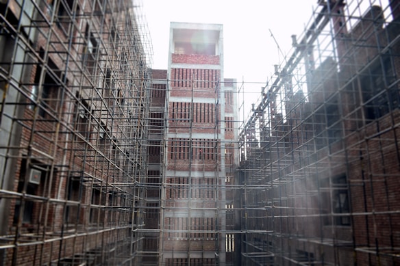 A construction site featuring tall structures covered in scaffolding, with exposed brickwork on the buildings. The scaffolds form a grid pattern around the buildings. The scene appears industrial and shows early stages of development.