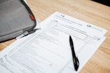 A tax form labeled Form 1120 sits on a wooden table. A black pen rests on the form, and a gray zippered pouch is partially visible beside it.
