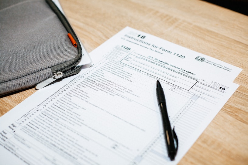 A tax form labeled Form 1120 sits on a wooden table. A black pen rests on the form, and a gray zippered pouch is partially visible beside it.